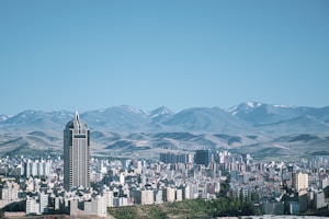 A sprawling cityscape featuring numerous modern buildings with a prominent skyscraper in the foreground. The backdrop consists of expansive mountain ranges with patches of snow. The color palette includes various shades of blue, gray, and earthy tones, giving a serene and expansive feel.
