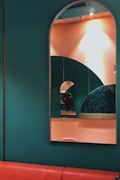 A series of mirrors reflecting each other are mounted on a wall painted in dark green and peach colors. The largest mirror has an arched top, creating a layered geometric effect with the reflection of smaller mirrors. The wall pattern includes an interesting design with circular shapes and different textures, adding depth to the composition. A red cushioned bench runs along the bottom of the wall.