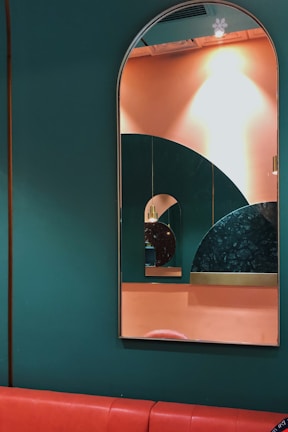 A series of mirrors reflecting each other are mounted on a wall painted in dark green and peach colors. The largest mirror has an arched top, creating a layered geometric effect with the reflection of smaller mirrors. The wall pattern includes an interesting design with circular shapes and different textures, adding depth to the composition. A red cushioned bench runs along the bottom of the wall.