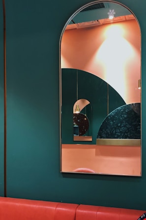 A series of mirrors reflecting each other are mounted on a wall painted in dark green and peach colors. The largest mirror has an arched top, creating a layered geometric effect with the reflection of smaller mirrors. The wall pattern includes an interesting design with circular shapes and different textures, adding depth to the composition. A red cushioned bench runs along the bottom of the wall.