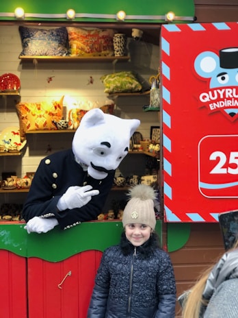 A child wearing a winter jacket and beanie poses in front of a colorful stall that displays various decorative items like pillows and ceramics. Next to the child, a person in a large white cat costume with a friendly expression is gesturing in a playful manner. The stall has vibrant signage with bold colors and patterns.
