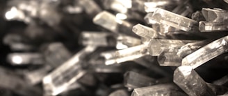 A close-up view of numerous transparent, elongated crystal formations tightly packed together. The crystals have sharp edges and reflective surfaces, capturing various shades of light.