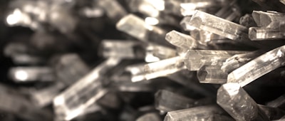 A close-up view of numerous transparent, elongated crystal formations tightly packed together. The crystals have sharp edges and reflective surfaces, capturing various shades of light.