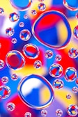 A vibrant collection of water droplets rests on a colorful background, creating a kaleidoscopic effect. The droplets magnify and distort the backdrop, composed of bold hues such as red, blue, yellow, and purple, adding depth and complexity to the visual experience.