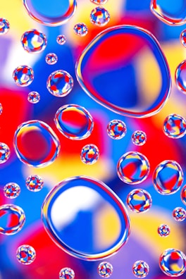 A vibrant collection of water droplets rests on a colorful background, creating a kaleidoscopic effect. The droplets magnify and distort the backdrop, composed of bold hues such as red, blue, yellow, and purple, adding depth and complexity to the visual experience.