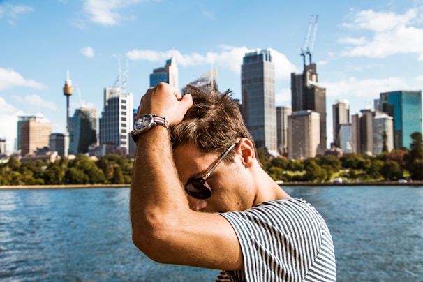 A stylish young man adjusting his sunglasses with a city skyline in the background.