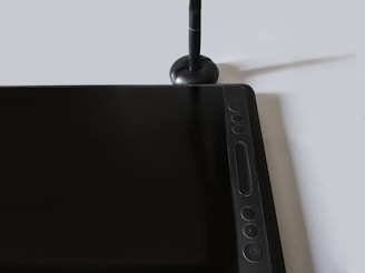 A digital drawing tablet with a stylus rests on a flat surface. The tablet features several function buttons and a sleek design. The stylus stands upright in a stylus holder next to the tablet.