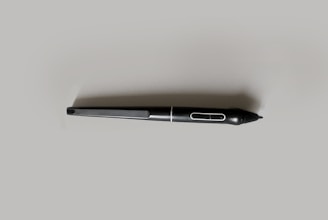A black digital stylus or pen with a streamlined design, featuring a button near the tip and a smooth, matte finish. It is placed on a light gray background, emphasizing its sleek form.
