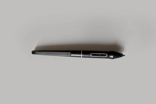A black digital stylus or pen with a streamlined design, featuring a button near the tip and a smooth, matte finish. It is placed on a light gray background, emphasizing its sleek form.