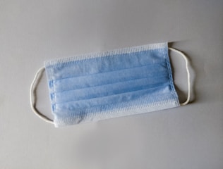 A disposable blue surgical mask with white ear loops lying flat on a neutral-colored surface.