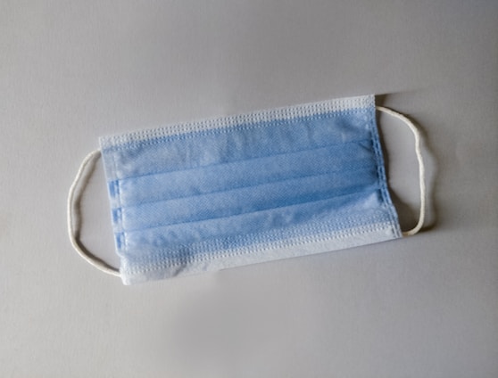 A disposable blue surgical mask with white ear loops lying flat on a neutral-colored surface.