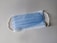 A disposable blue surgical mask with white ear loops lying flat on a neutral-colored surface.