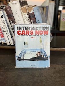 A collection of books is displayed, with one prominent book titled 'Intersection Cars Now' at the forefront. The book cover features a person driving a car, wearing sunglasses, set against a backdrop of blue and white designs. Surrounding books have titles about architecture and sculpture.