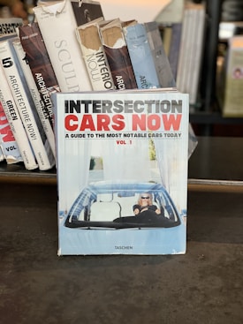 A collection of books is displayed, with one prominent book titled 'Intersection Cars Now' at the forefront. The book cover features a person driving a car, wearing sunglasses, set against a backdrop of blue and white designs. Surrounding books have titles about architecture and sculpture.