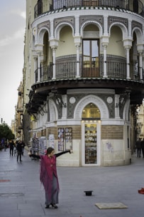 A street performer dressed in traditional attire dances in front of an ornately decorated building with arches and balconies. The building features intricate patterns and houses a souvenir shop at the ground level. Passersby can be seen walking along the street.