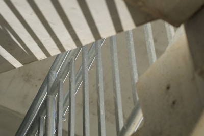 A close-up view of metal railings with geometric shadows cast on a concrete surface. The angles and lines create an abstract pattern, highlighting the industrial design.