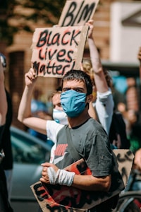 A person wearing a blue face mask and holding a sign participates in a protest. Other participants in the background also hold signs with messages like 'Black Lives Matter' and 'BLM'. The scene is outdoors, with a focus on activism and social justice.