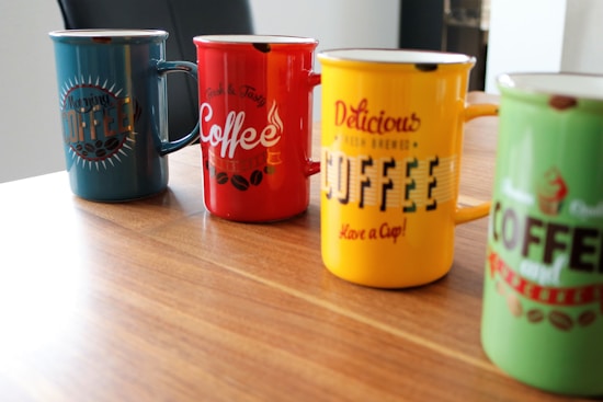 Four colorful coffee mugs are arranged on a wooden table, each displaying different typography related to coffee. The mugs are in shades of blue, red, yellow, and green, with text and decorative elements that convey a lively and vibrant atmosphere.