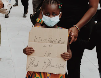 A young child wearing a colorful traditional dress holds a handwritten sign with the words 'It's a hard knock life I'm Black & Beautiful'. The child is wearing a face mask and is accompanied by an adult. Other people in the background are also wearing masks, suggesting a protest or public gathering. The scene takes place on a paved surface.
