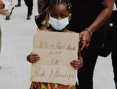 A young child wearing a colorful traditional dress holds a handwritten sign with the words 'It's a hard knock life I'm Black & Beautiful'. The child is wearing a face mask and is accompanied by an adult. Other people in the background are also wearing masks, suggesting a protest or public gathering. The scene takes place on a paved surface.