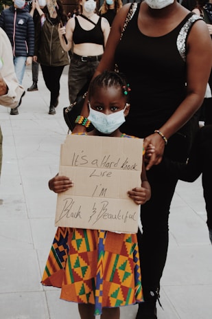 A young child wearing a colorful traditional dress holds a handwritten sign with the words 'It's a hard knock life I'm Black & Beautiful'. The child is wearing a face mask and is accompanied by an adult. Other people in the background are also wearing masks, suggesting a protest or public gathering. The scene takes place on a paved surface.