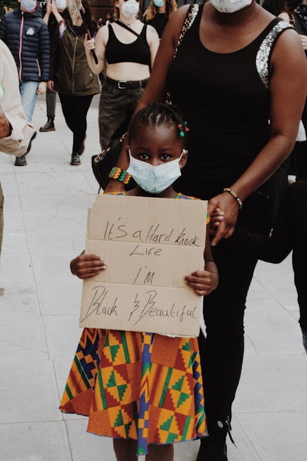 A young child wearing a colorful traditional dress holds a handwritten sign with the words 'It's a hard knock life I'm Black & Beautiful'. The child is wearing a face mask and is accompanied by an adult. Other people in the background are also wearing masks, suggesting a protest or public gathering. The scene takes place on a paved surface.