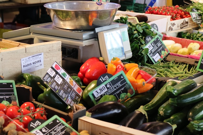 A vibrant market stall laden with a variety of fresh vegetables including red bell peppers, yellow bell peppers, green bell peppers, zucchini, eggplant, green beans, and tomatoes. There is a weighing scale located in the middle of the stall with a small digital display screen. Different price tags are visible, displaying prices per kilogram for various vegetables.