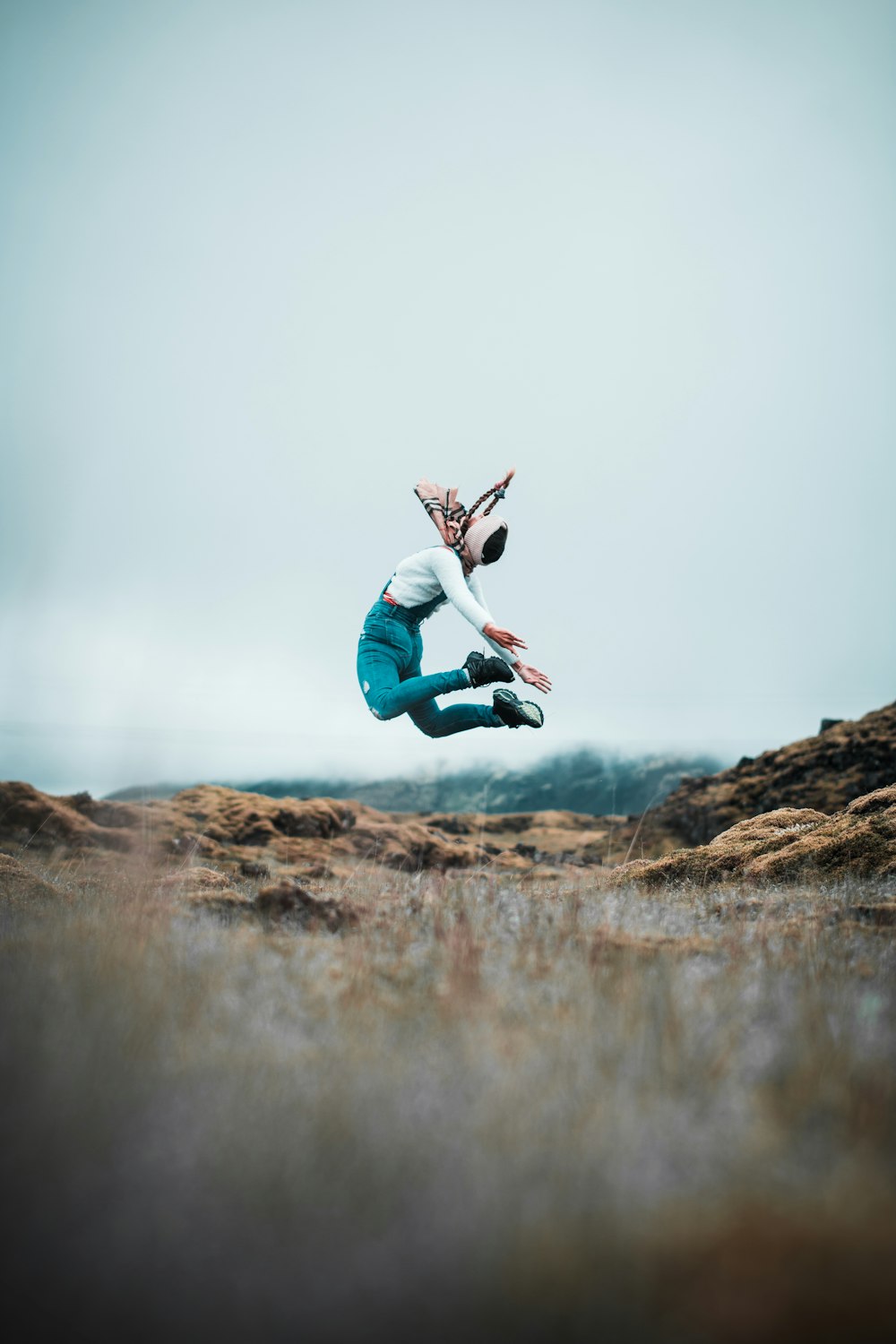 woman in white long sleeve shirt and blue denim jeans jumping on brown rock during daytime