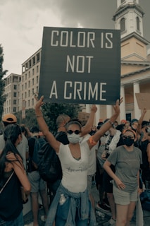 People are gathered in a protest, with a central focus on a person holding a large sign that reads 'Color is Not a Crime'. The crowd wears casual clothing and masks, suggesting a demonstration during a pandemic. The background includes urban architecture, such as a building and some trees.