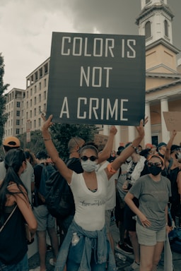 People are gathered in a protest, with a central focus on a person holding a large sign that reads 'Color is Not a Crime'. The crowd wears casual clothing and masks, suggesting a demonstration during a pandemic. The background includes urban architecture, such as a building and some trees.