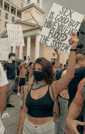 People are gathered in a protest, holding signs with bold messages. One sign prominently visible reads, 'Sex is good, but have you ever fucked the system.' Participants are wearing masks, indicating health precautions. The background includes a building with columns.