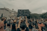 A large group of people is gathered in a city setting, some wearing face masks. A person in the foreground holds a sign reading 'No Justice No Peace' above their head. The crowd is diverse, and many are holding various signs. Buildings and trees are visible in the background under a cloudy sky.