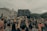 A large group of people is gathered in a city setting, some wearing face masks. A person in the foreground holds a sign reading 'No Justice No Peace' above their head. The crowd is diverse, and many are holding various signs. Buildings and trees are visible in the background under a cloudy sky.