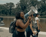 A group of people is walking near a body of water, with one woman holding a megaphone and appearing to speak passionately. She is accompanied by others who seem engaged and are holding signs. The setting includes trees and a clear sky.