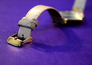 A close-up view of a leather watch strap with a metallic buckle resting on a rich purple surface. The buckle has a shiny gold finish, and the texture of the leather is clearly visible, giving a sense of elegance and craftsmanship.