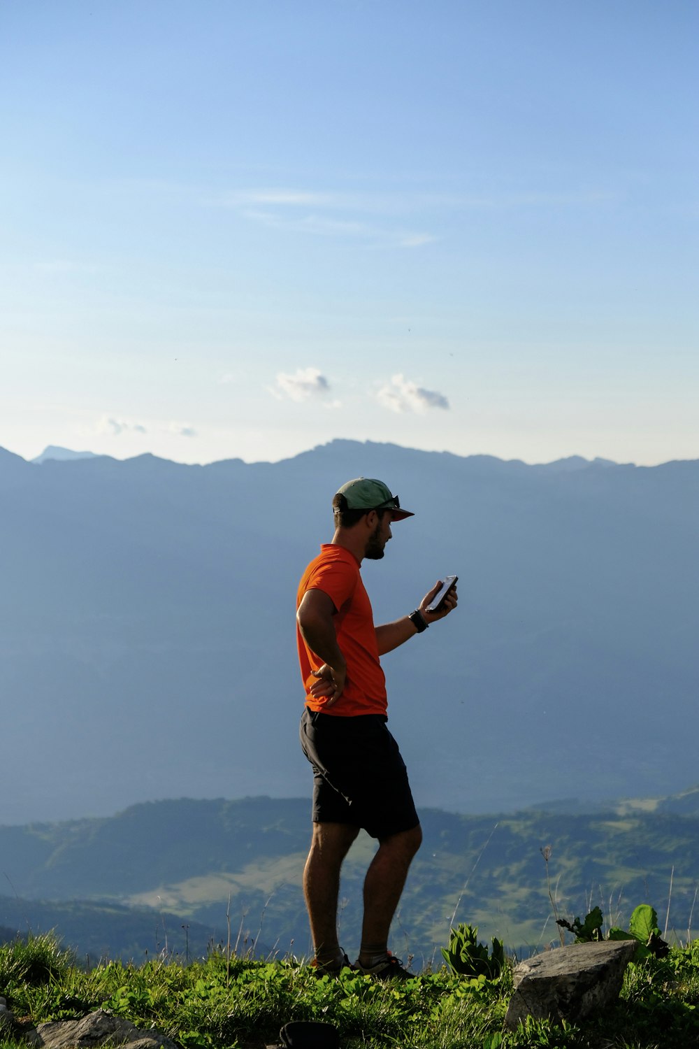 man in orange tank top and black shorts holding camera standing on top of mountain during