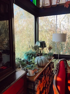 A cozy sunlit corner features a small wooden table adorned with a variety of potted plants and a decorative white pumpkin. Below the table, stacks of firewood add a rustic touch. Natural light streams in through large windows, creating a warm ambiance. A red-upholstered chair sits nearby, next to a white lamp, enhancing the cozy setting.