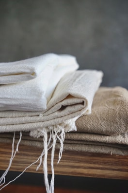 A stack of folded fabric pieces is placed on a wooden surface. The fabrics consist of various natural tones, with some having frayed edges suggesting a handmade or rustic quality. The backdrop is a plain, muted wall that complements the earthy colors of the cloth.