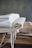 A stack of folded fabric pieces is placed on a wooden surface. The fabrics consist of various natural tones, with some having frayed edges suggesting a handmade or rustic quality. The backdrop is a plain, muted wall that complements the earthy colors of the cloth.