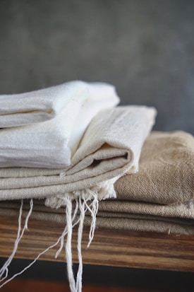 A stack of folded fabric pieces is placed on a wooden surface. The fabrics consist of various natural tones, with some having frayed edges suggesting a handmade or rustic quality. The backdrop is a plain, muted wall that complements the earthy colors of the cloth.