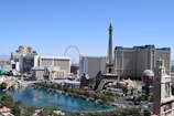 A vibrant cityscape featuring iconic landmarks such as a replica of the Eiffel Tower, high-rise buildings, and a large Ferris wheel. A serene turquoise water body runs along the strip, bordered by lush greenery and contemporary architecture.