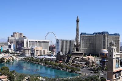A vibrant cityscape featuring iconic landmarks such as a replica of the Eiffel Tower, high-rise buildings, and a large Ferris wheel. A serene turquoise water body runs along the strip, bordered by lush greenery and contemporary architecture.