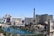 A vibrant cityscape featuring iconic landmarks such as a replica of the Eiffel Tower, high-rise buildings, and a large Ferris wheel. A serene turquoise water body runs along the strip, bordered by lush greenery and contemporary architecture.