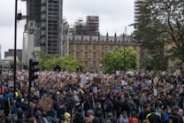 A large crowd of people gathered in a public square, many holding signs and banners advocating for social justice, with the recognizable architecture of a historical building in the background. The atmosphere is bustling, with individuals wearing masks and showing solidarity through their messages.