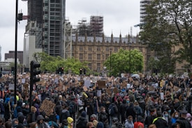 A large crowd of people gathered in a public square, many holding signs and banners advocating for social justice, with the recognizable architecture of a historical building in the background. The atmosphere is bustling, with individuals wearing masks and showing solidarity through their messages.