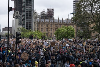 A large crowd of people gathered in a public square, many holding signs and banners advocating for social justice, with the recognizable architecture of a historical building in the background. The atmosphere is bustling, with individuals wearing masks and showing solidarity through their messages.