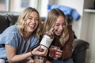 Two lively female hosts laughing together in a cozy podcast studio filled with colorful notes and coffee mugs.