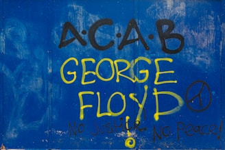 Graffiti featuring the text 'A.C.A.B' and 'George Floyd' is prominently displayed in large yellow and black letters against a blue backdrop. The phrases 'No Justice, No Peace!' accompany the text, along with a peace symbol.