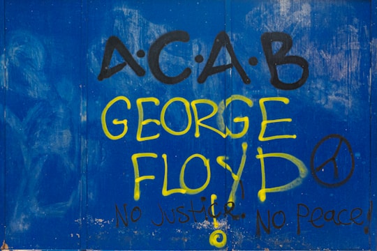 Graffiti featuring the text 'A.C.A.B' and 'George Floyd' is prominently displayed in large yellow and black letters against a blue backdrop. The phrases 'No Justice, No Peace!' accompany the text, along with a peace symbol.