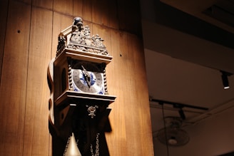 An ornate wall clock with intricate details is mounted on a wooden panel. The clock face features Roman numerals and decorative blue accents. A pendulum hangs below the clock, adding to its vintage appeal.
